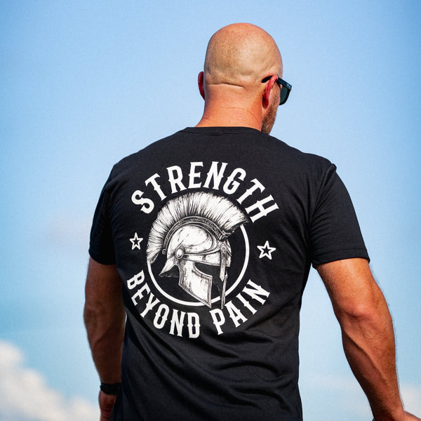 Man wearing a black t-shirt with 'Strength Beyond Pain' design against a blue sky.